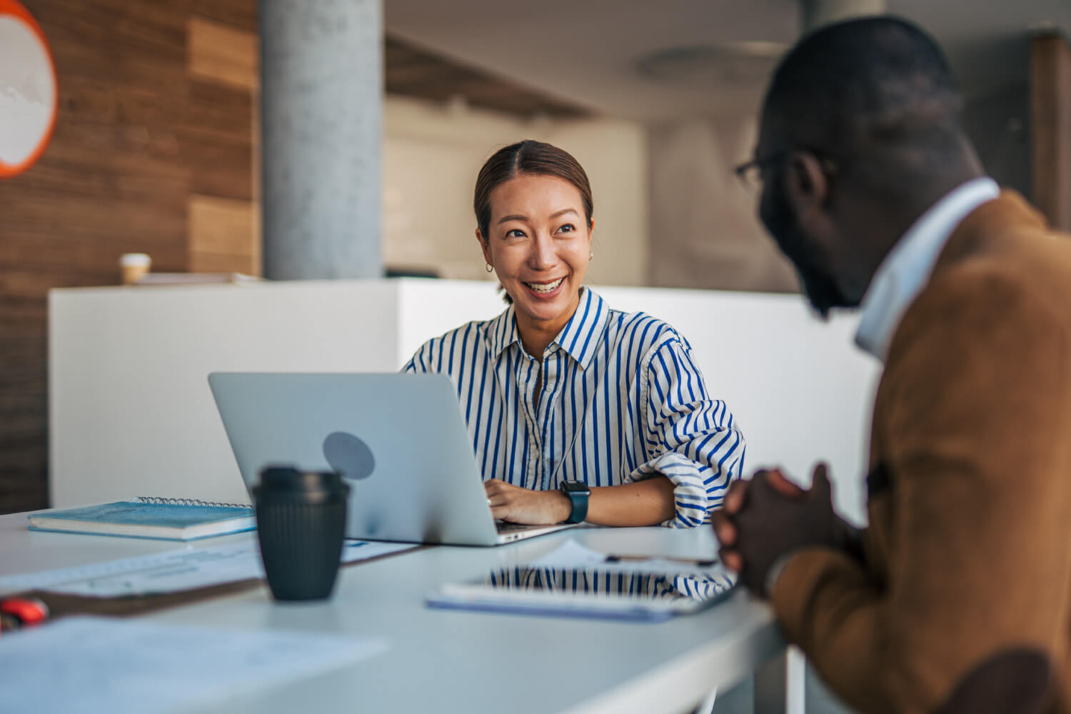 A woman smiling and talking with a colleague across a table in a modern office. She has a laptop open in front of her, and they appear to be having a friendly, productive conversation.