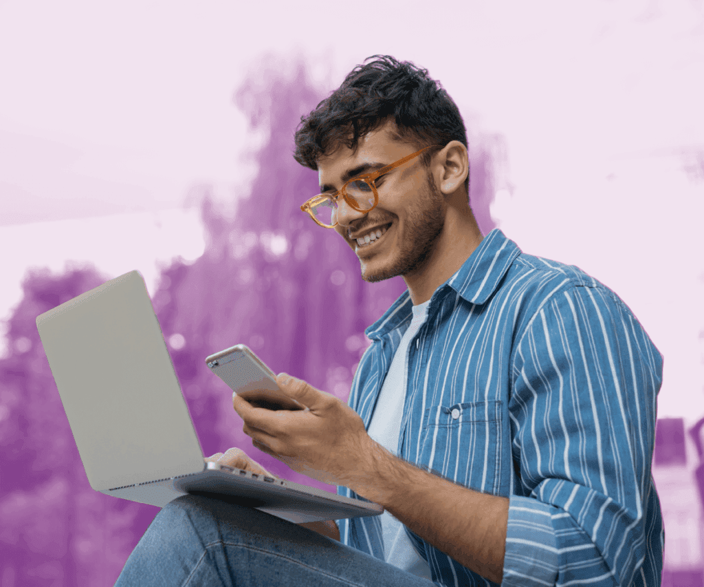 A smiling person sits outdoors with a laptop on their knees and a phone in hand, appearing engaged while multitasking between devices. They wear a striped blue shirt and glasses. The background is softly blurred with a purple gradient overlay, creating an uplifting, modern feel.