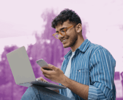 A smiling person sits outdoors with a laptop on their knees and a phone in hand, appearing engaged while multitasking between devices. They wear a striped blue shirt and glasses. The background is softly blurred with a purple gradient overlay, creating an uplifting, modern feel.