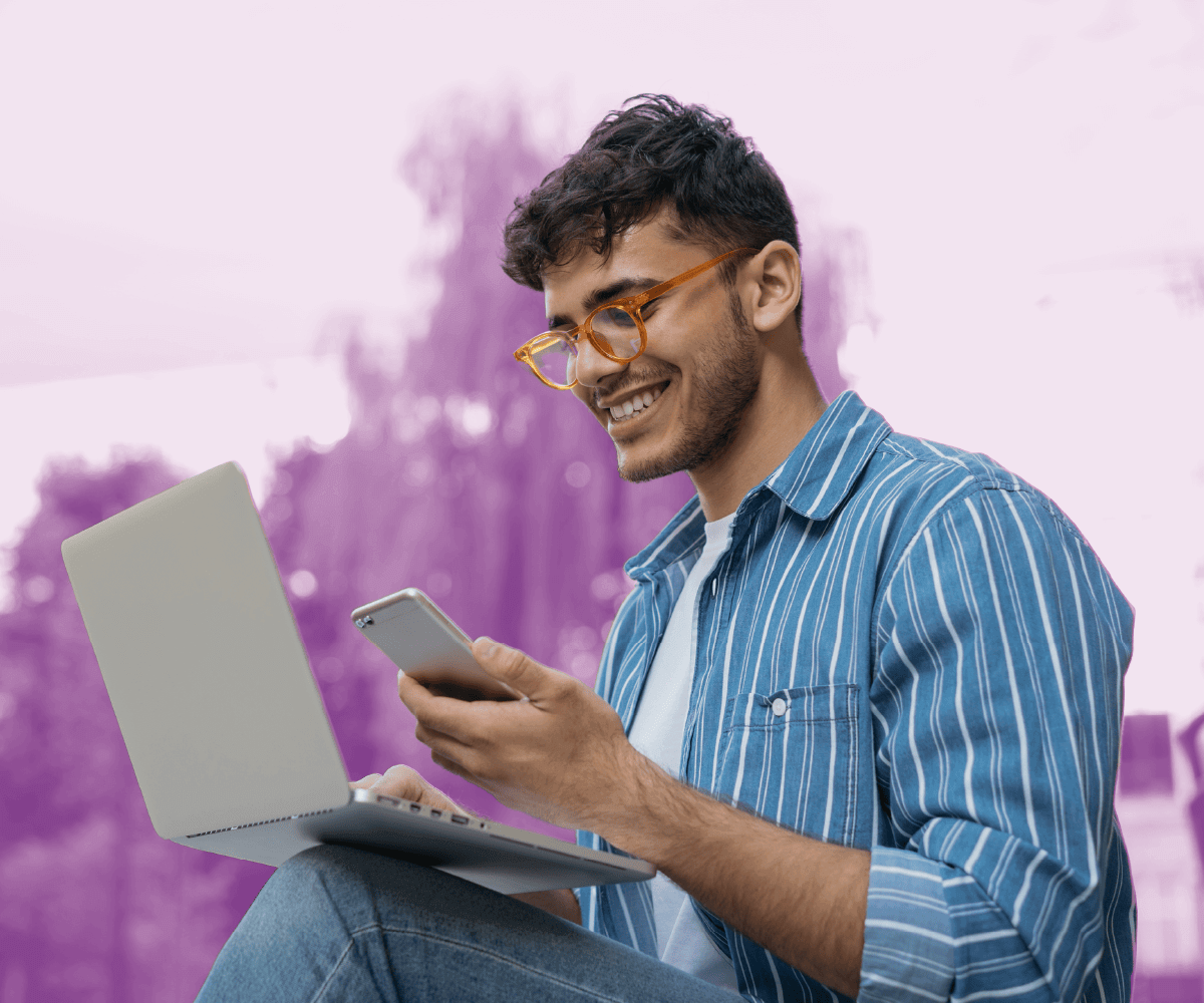 A smiling person sits outdoors with a laptop on their knees and a phone in hand, appearing engaged while multitasking between devices. They wear a striped blue shirt and glasses. The background is softly blurred with a purple gradient overlay, creating an uplifting, modern feel.