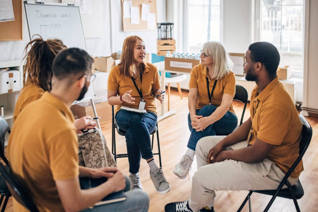 A group of five volunteers wearing matching mustard-colored shirts sit in a circle having a discussion inside a bright community room. One woman speaks while others listen attentively, with a whiteboard behind them labeled “Donation Goals.” Boxes and bottled water are visible in the background, suggesting a planning meeting for a community or charity initiative.