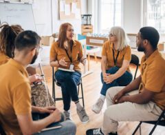 A group of five volunteers wearing matching mustard-colored shirts sit in a circle having a discussion inside a bright community room. One woman speaks while others listen attentively, with a whiteboard behind them labeled “Donation Goals.” Boxes and bottled water are visible in the background, suggesting a planning meeting for a community or charity initiative.