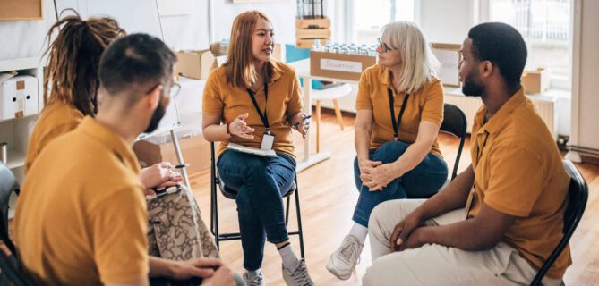 A group of five volunteers wearing matching mustard-colored shirts sit in a circle having a discussion inside a bright community room. One woman speaks while others listen attentively, with a whiteboard behind them labeled “Donation Goals.” Boxes and bottled water are visible in the background, suggesting a planning meeting for a community or charity initiative.
