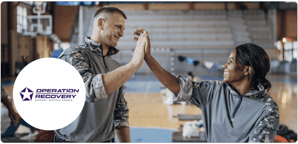 Two volunteers wearing gray camouflage uniforms smile and give each other a high-five in a gymnasium. A white circle on the left displays the purple Operation Recovery logo with the tagline “Support. Sustain. Enable.”