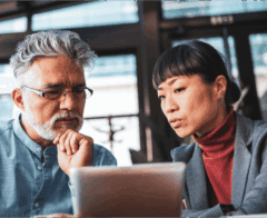 A man and woman sit together in a modern office, focusing intently on a tablet screen. The man rests his chin on his hand while the woman points or gestures toward the tablet, suggesting they are reviewing or discussing information related to AI or data analysis.