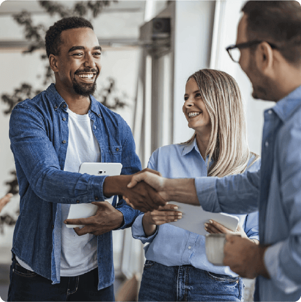 Three colleagues stand in a bright office space, smiling and shaking hands in greeting. One man holds a tablet while another holds a coffee cup, representing partnership and successful collaboration.