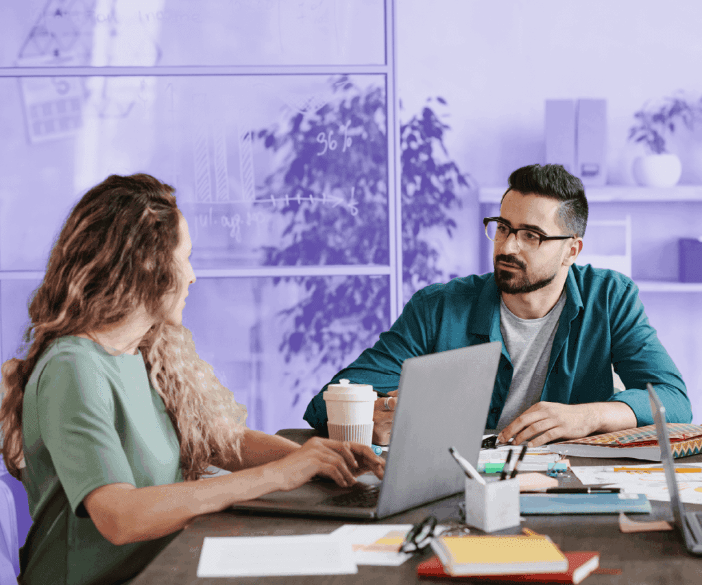 Two colleagues are sitting at a desk in a modern office, discussing work while using laptops. The man in glasses listens attentively to the woman as they collaborate. Papers, pens, and a coffee cup are on the table, and a glass wall with charts drawn on it is visible in the background.