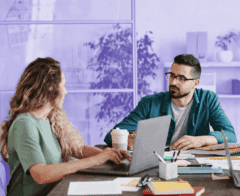 Two colleagues are sitting at a desk in a modern office, discussing work while using laptops. The man in glasses listens attentively to the woman as they collaborate. Papers, pens, and a coffee cup are on the table, and a glass wall with charts drawn on it is visible in the background.