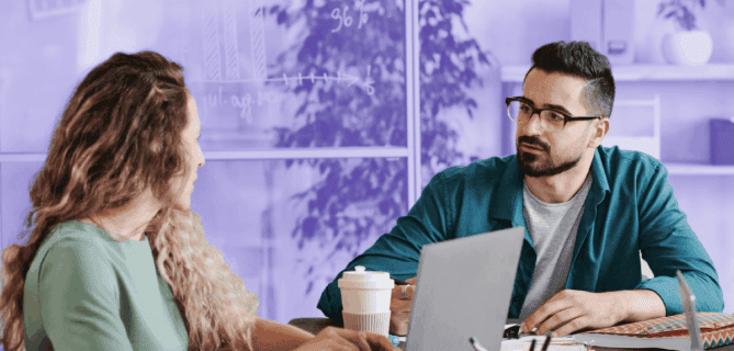 Two colleagues are sitting at a desk in a modern office, discussing work while using laptops. The man in glasses listens attentively to the woman as they collaborate. Papers, pens, and a coffee cup are on the table, and a glass wall with charts drawn on it is visible in the background.