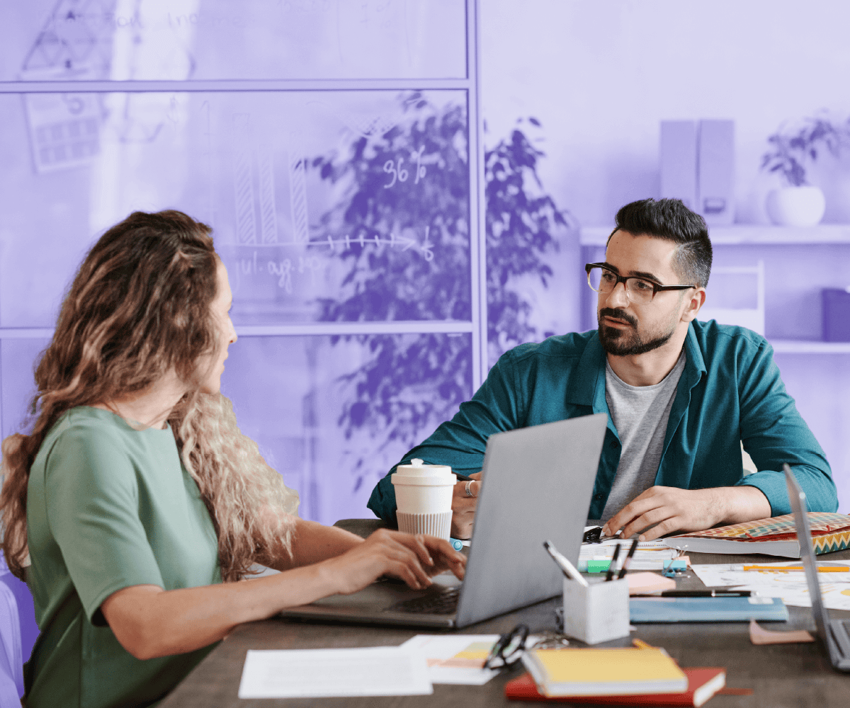 Two colleagues are sitting at a desk in a modern office, discussing work while using laptops. The man in glasses listens attentively to the woman as they collaborate. Papers, pens, and a coffee cup are on the table, and a glass wall with charts drawn on it is visible in the background.