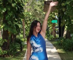 Cassandra Stone smiles and waves while walking along a tropical pathway surrounded by lush greenery.