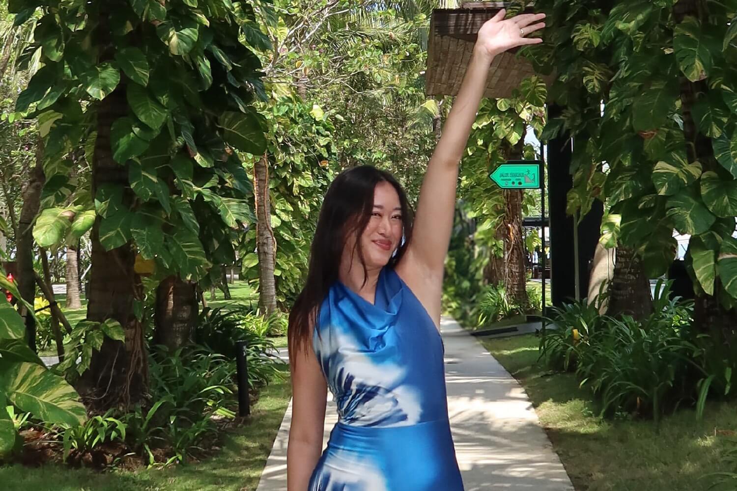 Cassandra Stone smiles and waves while walking along a tropical pathway surrounded by lush greenery.