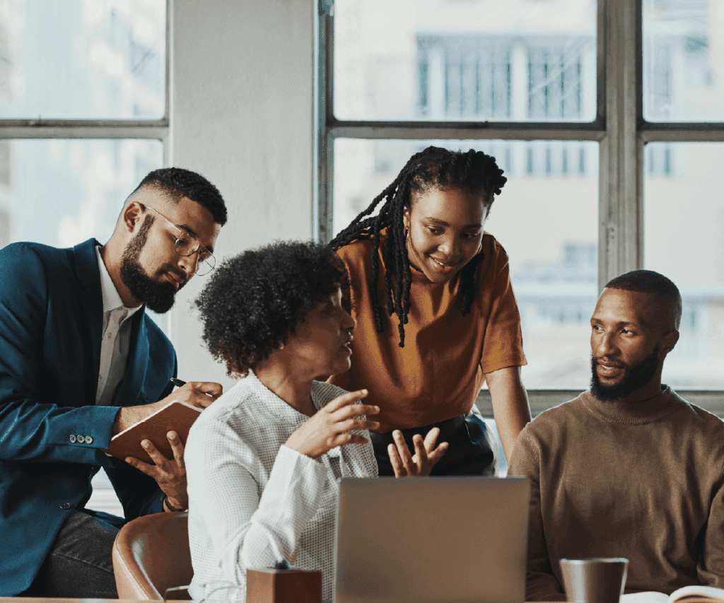 A group of four professionals in a bright office collaborates around a laptop. One woman speaks while others listen attentively, taking notes. The setting conveys teamwork, inclusion, and shared problem-solving.