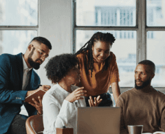 A group of four professionals in a bright office collaborates around a laptop. One woman speaks while others listen attentively, taking notes. The setting conveys teamwork, inclusion, and shared problem-solving.