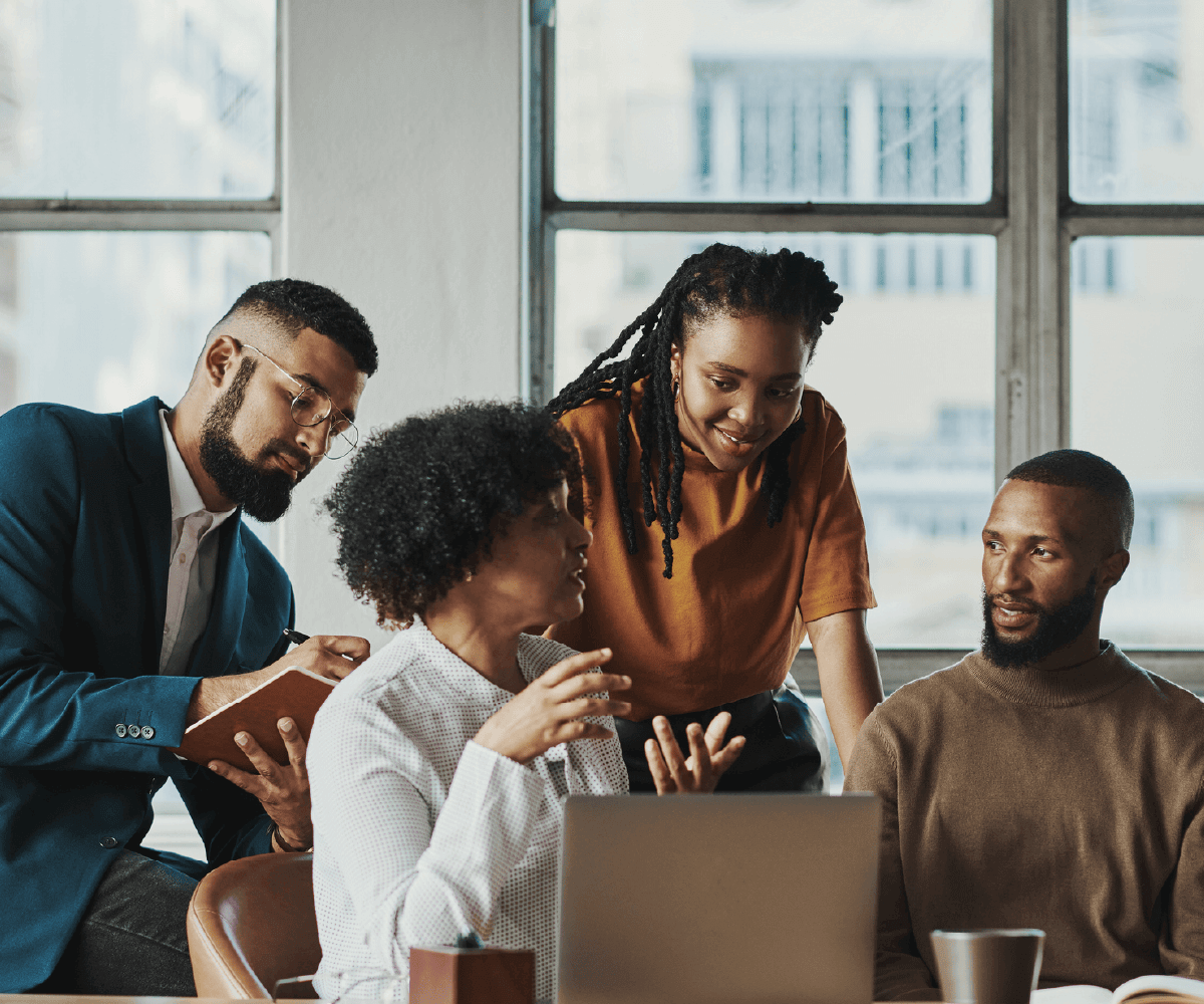 A group of four professionals in a bright office collaborates around a laptop. One woman speaks while others listen attentively, taking notes. The setting conveys teamwork, inclusion, and shared problem-solving.