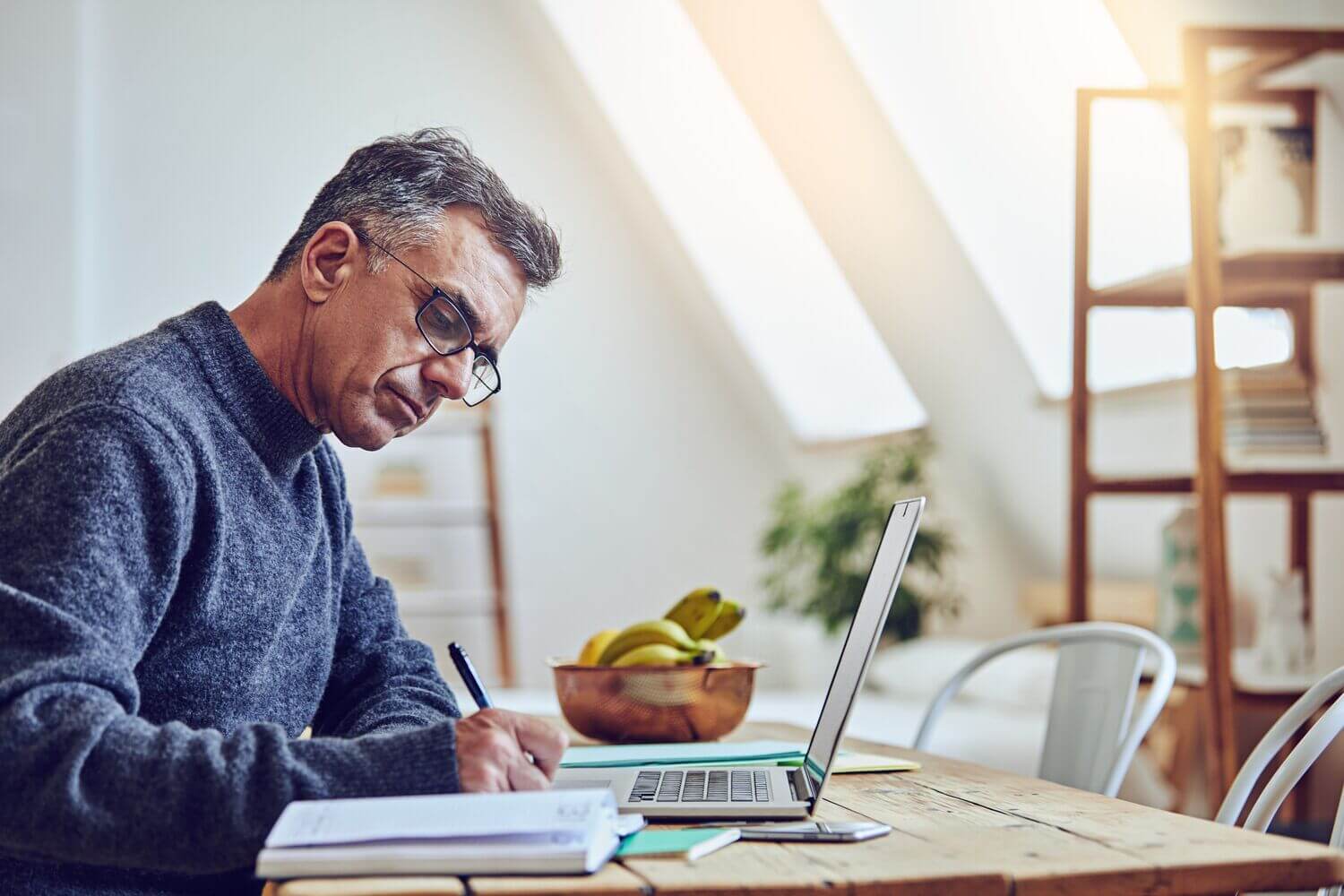 A man sits at a wooden table in a bright home office, focused on writing notes while working on his laptop. Sunlight filters through the windows, creating a calm, productive atmosphere—illustrating learning, reflection, and personal growth.