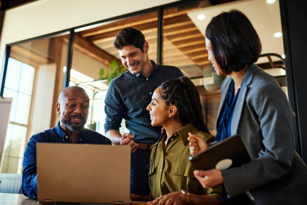 A diverse group of four professionals gathers around a laptop in a bright, modern office. They smile and engage in discussion, suggesting teamwork and collaboration. The image conveys partnership, communication, and productivity in a professional setting.