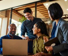A diverse group of four professionals gathers around a laptop in a bright, modern office. They smile and engage in discussion, suggesting teamwork and collaboration. The image conveys partnership, communication, and productivity in a professional setting.