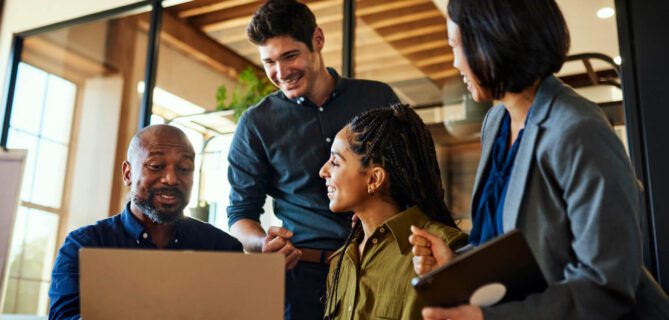 A diverse group of four professionals gathers around a laptop in a bright, modern office. They smile and engage in discussion, suggesting teamwork and collaboration. The image conveys partnership, communication, and productivity in a professional setting.