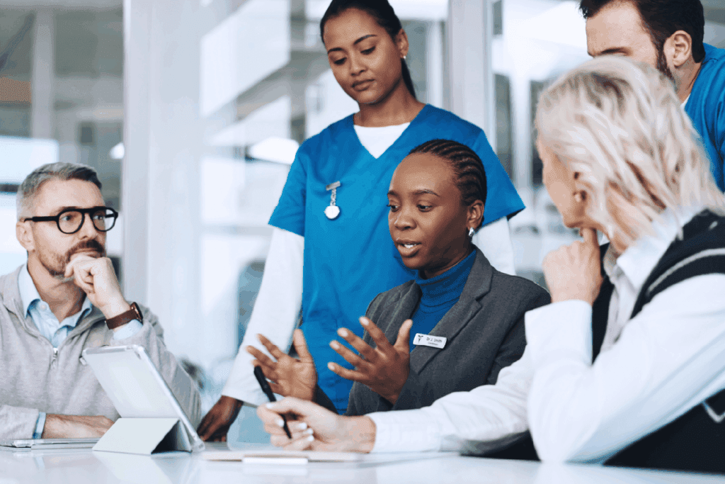 A diverse group of professionals, including healthcare workers in blue scrubs and others in business attire, gather around a conference table in discussion. A woman with a name badge speaks while others listen attentively, with tablets and documents in front of them. The scene conveys collaboration between medical and administrative teams in a professional setting.