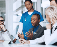 A diverse group of professionals, including healthcare workers in blue scrubs and others in business attire, gather around a conference table in discussion. A woman with a name badge speaks while others listen attentively, with tablets and documents in front of them. The scene conveys collaboration between medical and administrative teams in a professional setting.