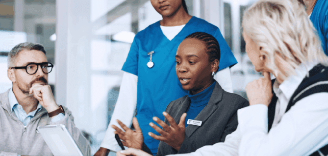 A diverse group of professionals, including healthcare workers in blue scrubs and others in business attire, gather around a conference table in discussion. A woman with a name badge speaks while others listen attentively, with tablets and documents in front of them. The scene conveys collaboration between medical and administrative teams in a professional setting.