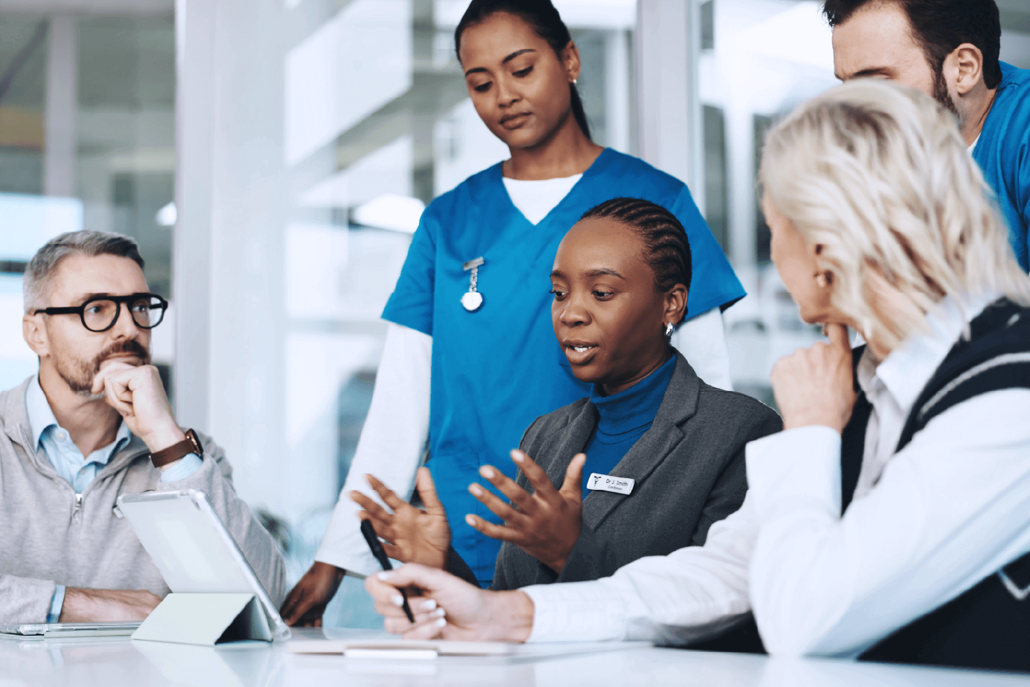 A diverse group of professionals, including healthcare workers in blue scrubs and others in business attire, gather around a conference table in discussion. A woman with a name badge speaks while others listen attentively, with tablets and documents in front of them. The scene conveys collaboration between medical and administrative teams in a professional setting.