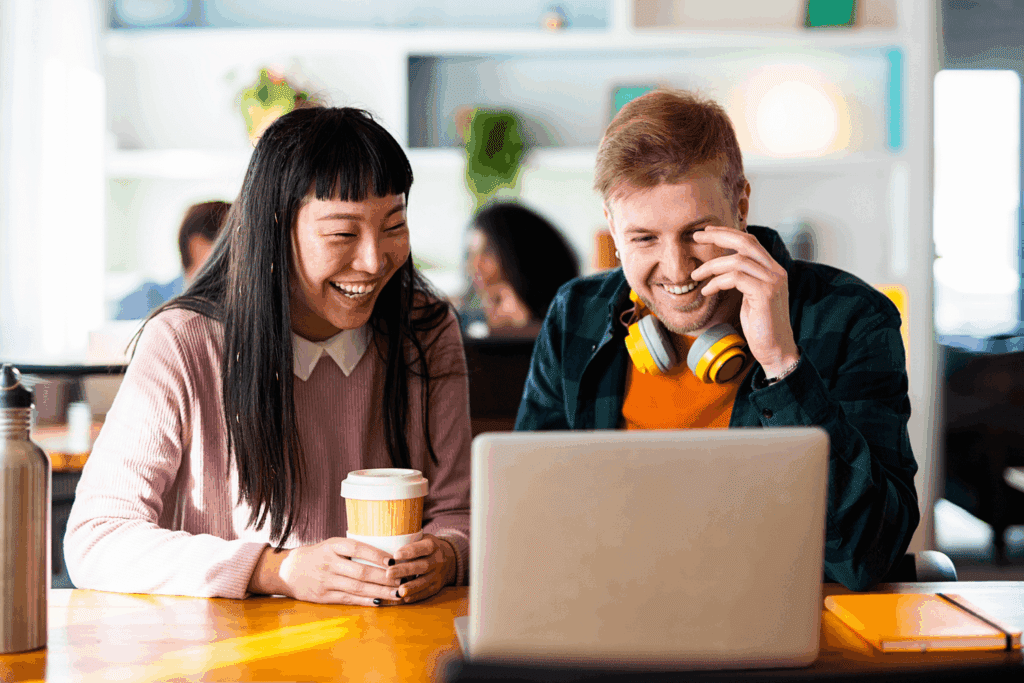 Two colleagues sitting at a table, smiling and looking at a laptop screen, representing collaboration and positive engagement in a shared workspace.