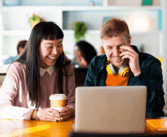 Two colleagues sitting at a table, smiling and looking at a laptop screen, representing collaboration and positive engagement in a shared workspace.