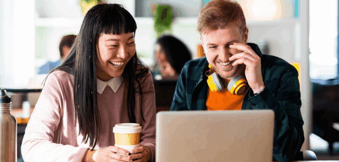 Two colleagues sitting at a table, smiling and looking at a laptop screen, representing collaboration and positive engagement in a shared workspace.