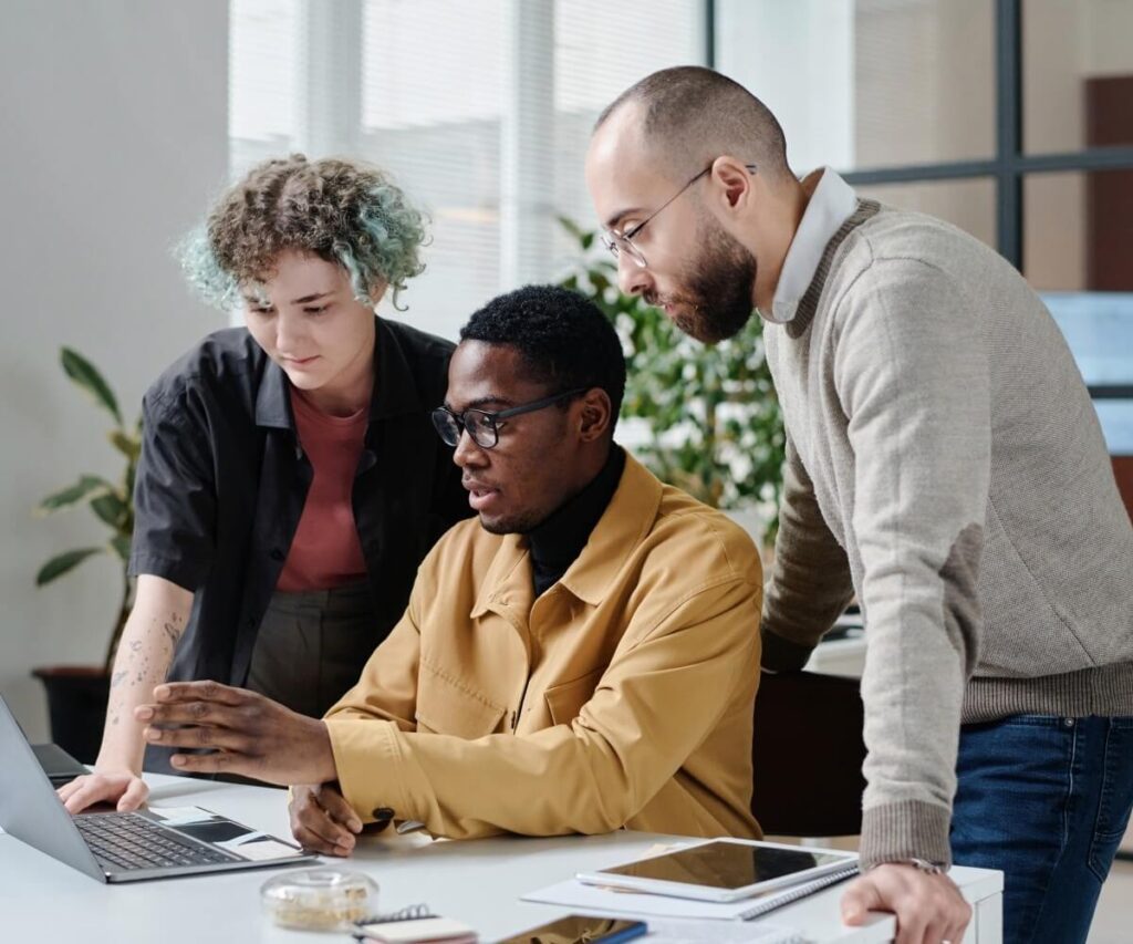 Three colleagues collaborate around a laptop at a desk, reviewing information together in a modern office.