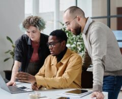 Three colleagues collaborate around a laptop at a desk, reviewing information together in a modern office.