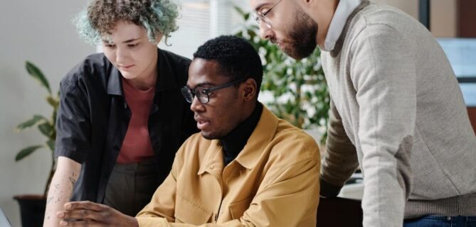 Three colleagues collaborate around a laptop at a desk, reviewing information together in a modern office.