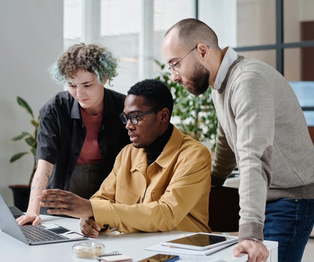 Three colleagues collaborate around a laptop at a desk, reviewing information together in a modern office.