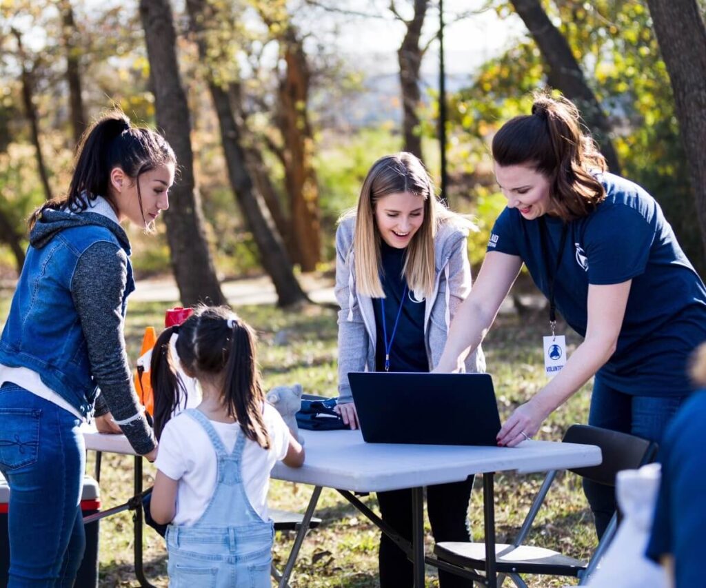 Volunteers gather around a table outdoors, using a laptop to coordinate activities during a community event.