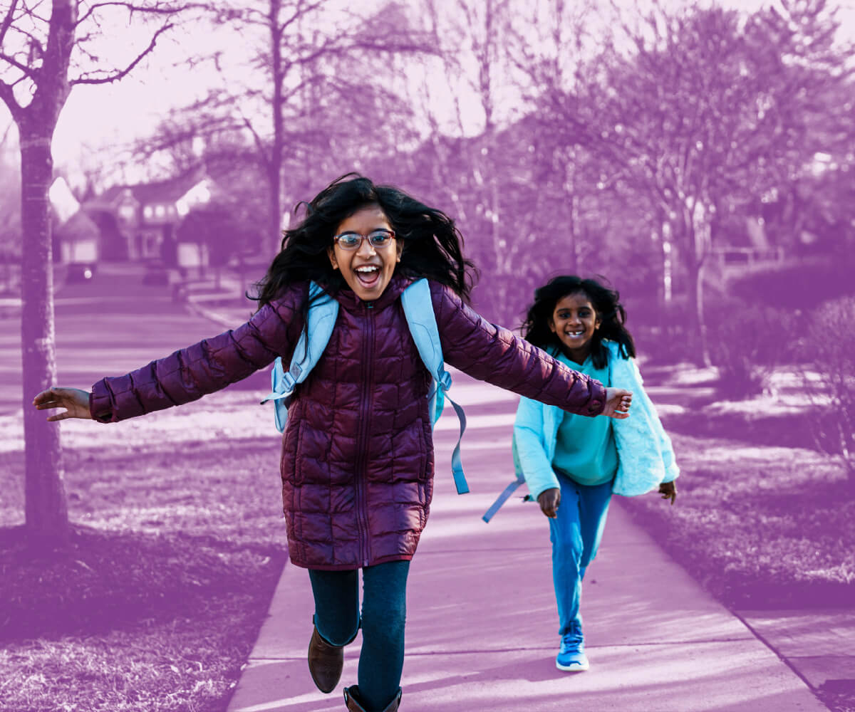 Two children wearing backpacks run joyfully along a paved path in a park, representing youth safety and community wellbeing.