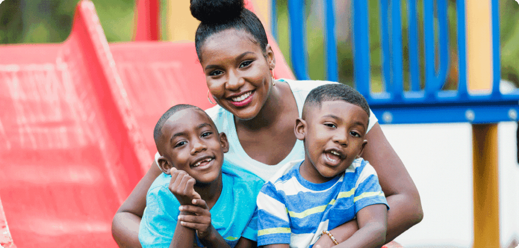 A smiling woman sits on playground equipment with two young children, symbolizing family support and community services.