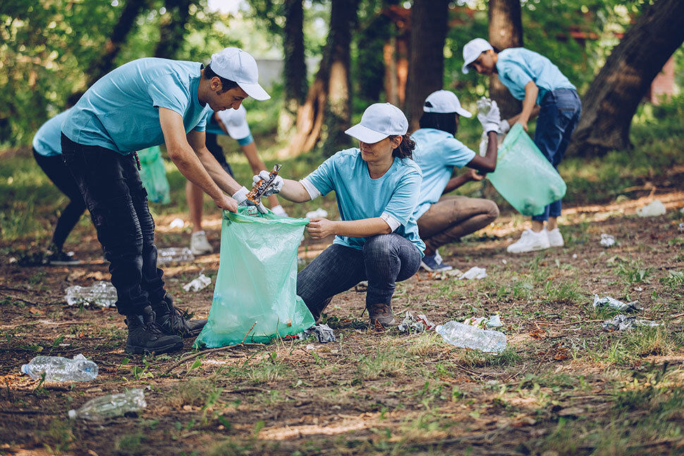A group of volunteers wearing matching shirts collect litter in a wooded park during a community cleanup effort.