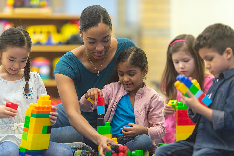 An adult guides young children as they build with colorful blocks in a classroom or childcare setting.