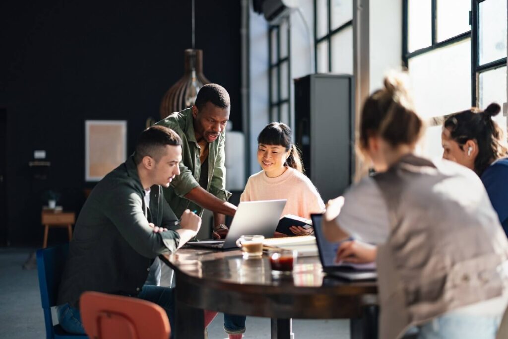 A small group of professionals collaborate around a table, reviewing information together on a laptop in a bright workspace.