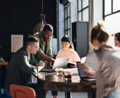 A small group of professionals collaborate around a table, reviewing information together on a laptop in a bright workspace.