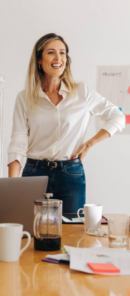 Smiling woman standing in a bright office beside a table with a laptop, coffee mugs, and papers, appearing to lead or present during a meeting.