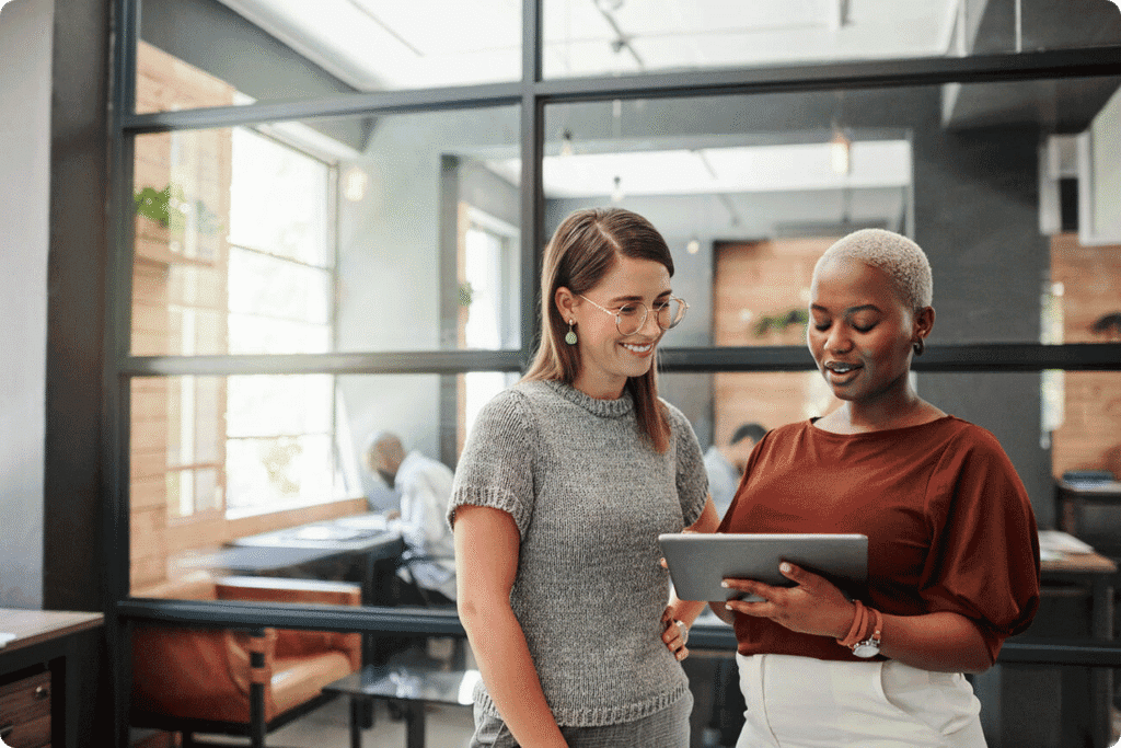 Two colleagues stand together in an office, looking at a tablet and talking.