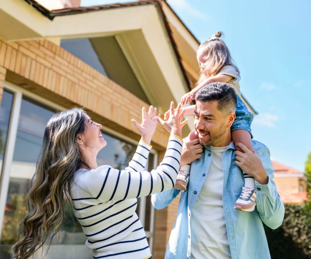 A family stands outside a home, smiling as a parent lifts a child onto their shoulders, representing housing stability and family support.