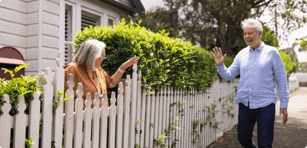 Two older neighbors smile and wave to each other across a white picket fence in a sunny residential neighborhood.