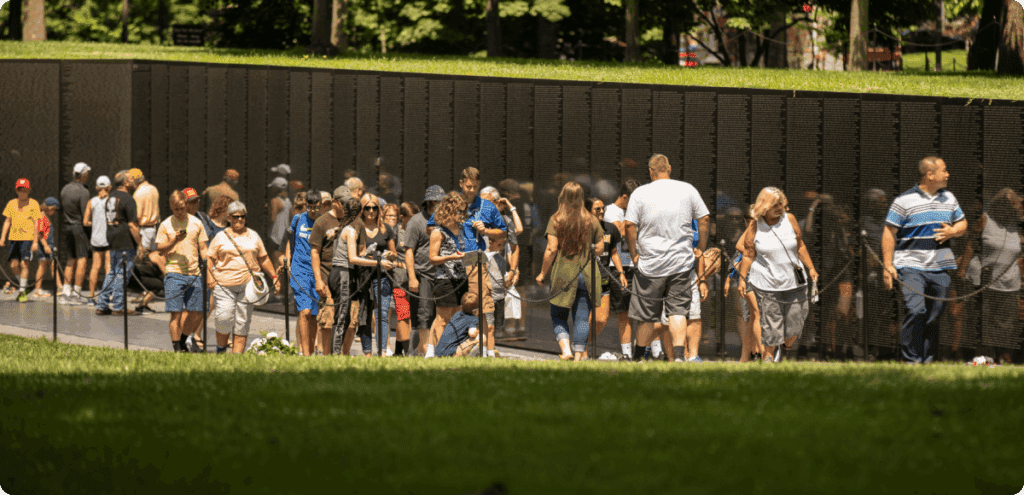 A large group of visitors gather along a memorial wall, reading inscriptions and walking through a tree-lined park.