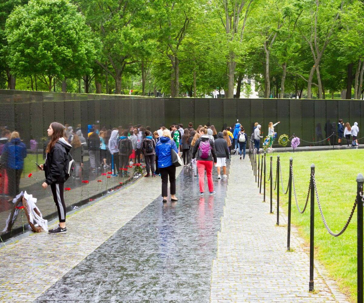 Visitors walk along a long black memorial wall outdoors, reading names and reflecting in a landscaped park setting.
