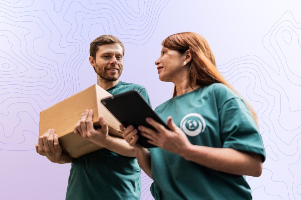 Two volunteers stand together reviewing a tablet while holding a box, representing coordinated work and operational readiness.