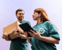 Two volunteers stand together reviewing a tablet while holding a box, representing coordinated work and operational readiness.