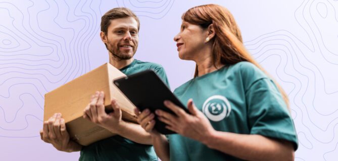 Two volunteers stand together reviewing a tablet while holding a box, representing coordinated work and operational readiness.